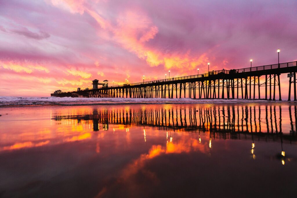 A stunning sunset over a pier with vibrant colors reflected in the wet sand below.
