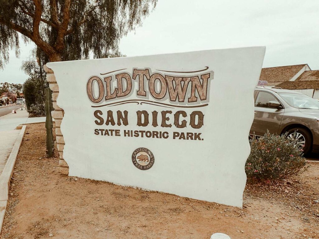 A large white sign at the entrance of Old Town San Diego State Historic Park with parked cars and trees nearby.