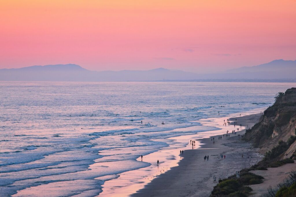 A serene beach with soft waves and silhouettes of people walking, under a pink and purple sunset sky.
