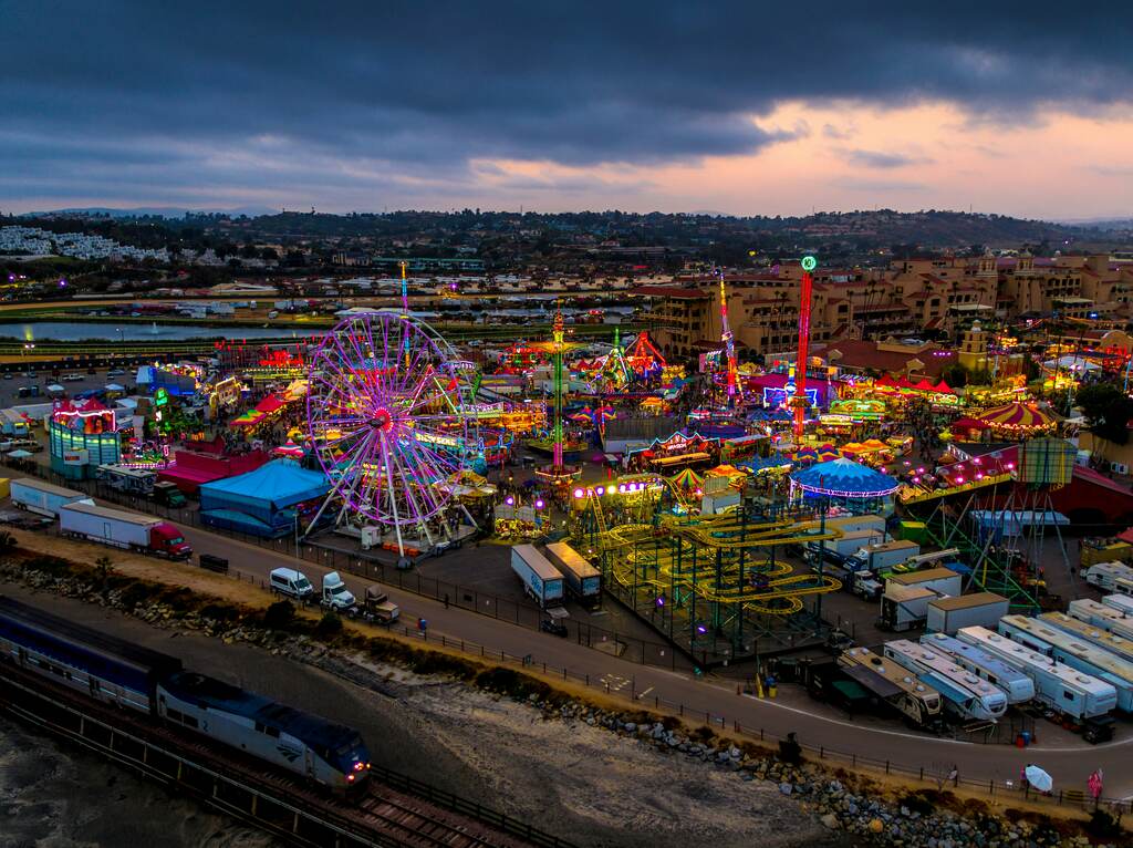 A vibrant aerial view of a carnival illuminated by colorful lights, with a ferris wheel and roller coaster rides at dusk.