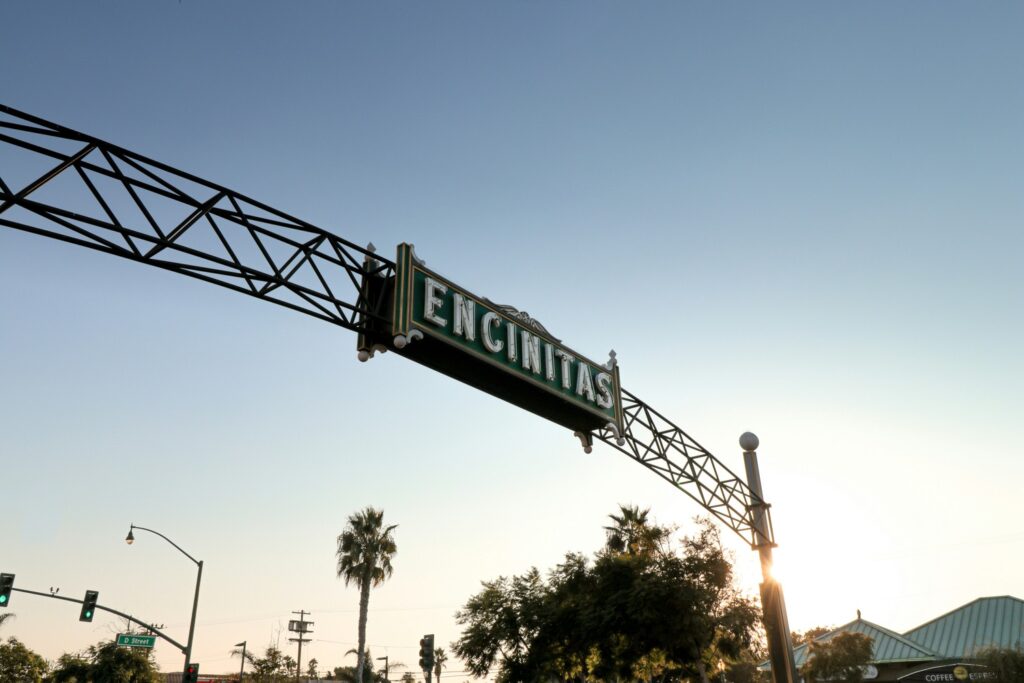 A metal archway sign reading "ENCINITAS" stretches across a street at sunset with palm trees and traffic lights in the background.
