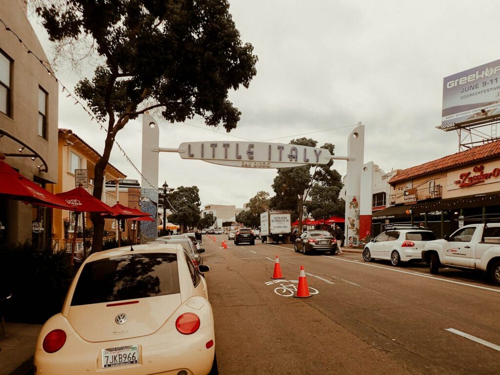 Street view of the entrance archway to Little Italy in San Diego with parked cars and outdoor seating under red umbrellas.
