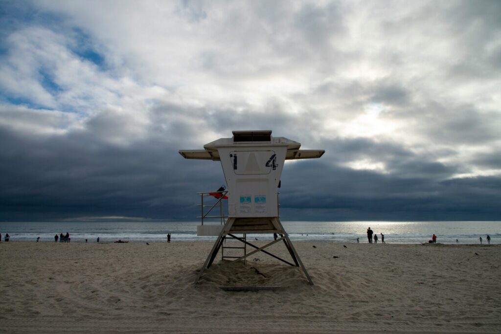 A lifeguard tower stands on a sandy beach under a dramatic cloudy sky with people near the shoreline.
