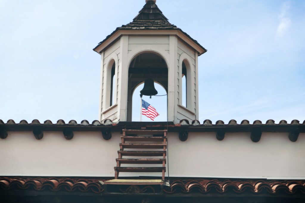 A bell tower with an American flag visible through the arch, set against a clear blue sky.