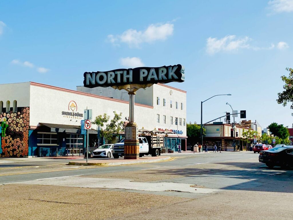 The image shows the iconic "North Park" sign in San Diego with local businesses and traffic in the background on a sunny day.