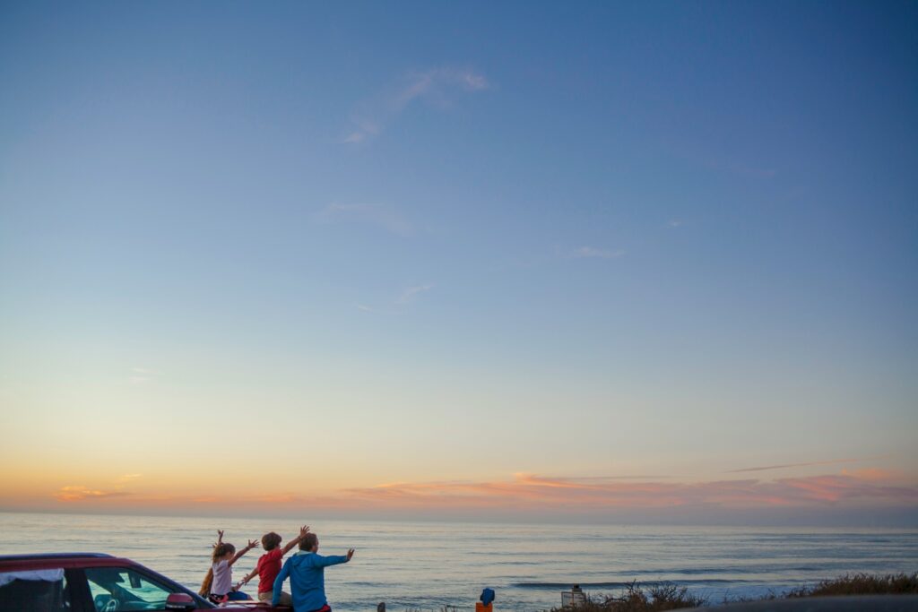 Three children waving toward the ocean at sunset, sitting on a car roof near the beach.
