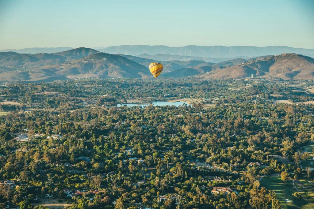 A yellow hot air balloon floats above the lush, green landscape of Rancho Santa Fe, California, with mountains in the background.