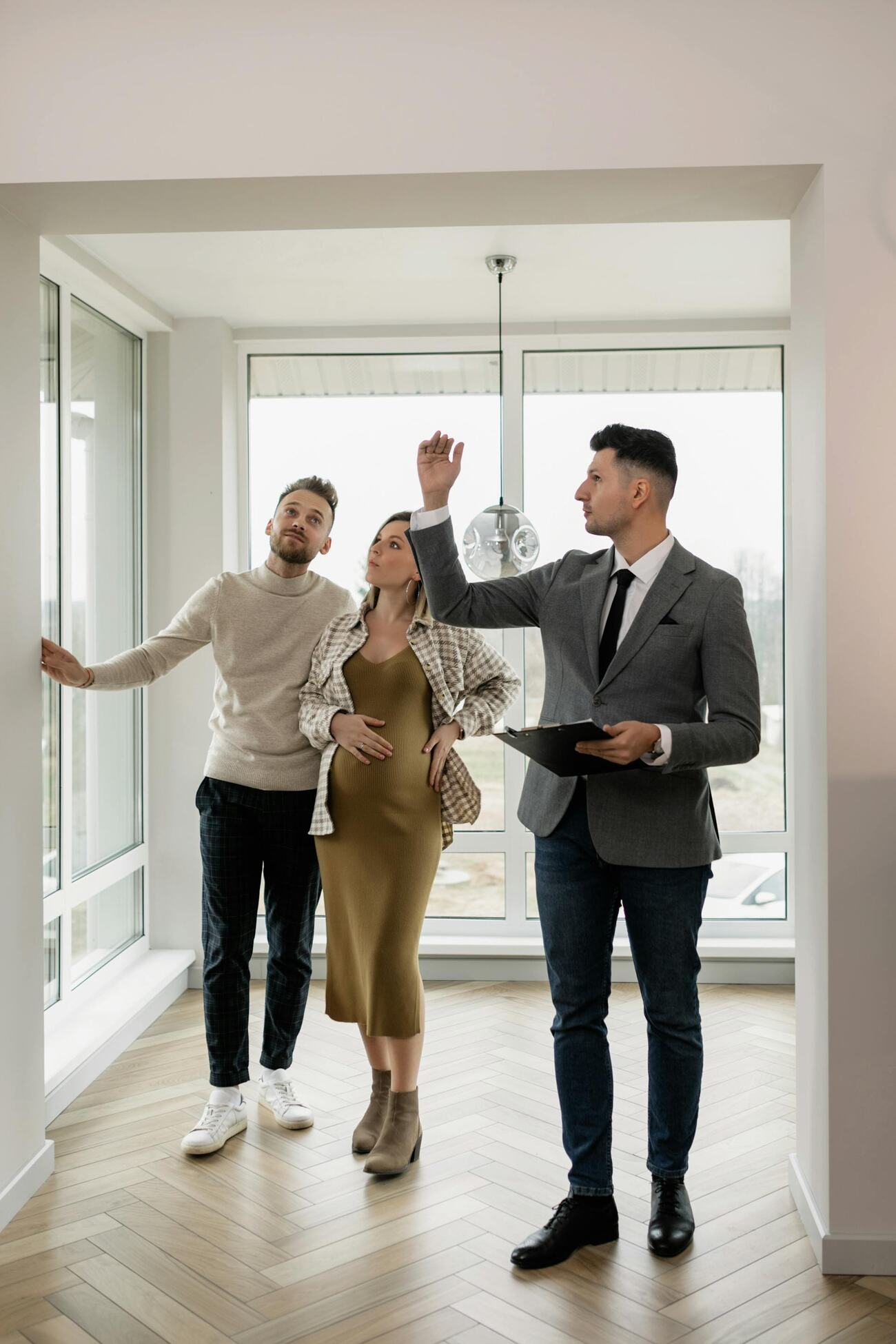 A man points to something while standing with a man and pregnant woman in an open room