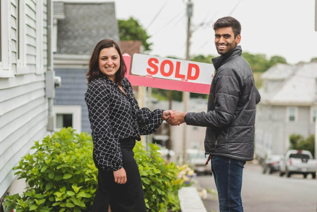 A man and woman shakes hands in front of a "Sold" sign, celebrating the successful sale of a property