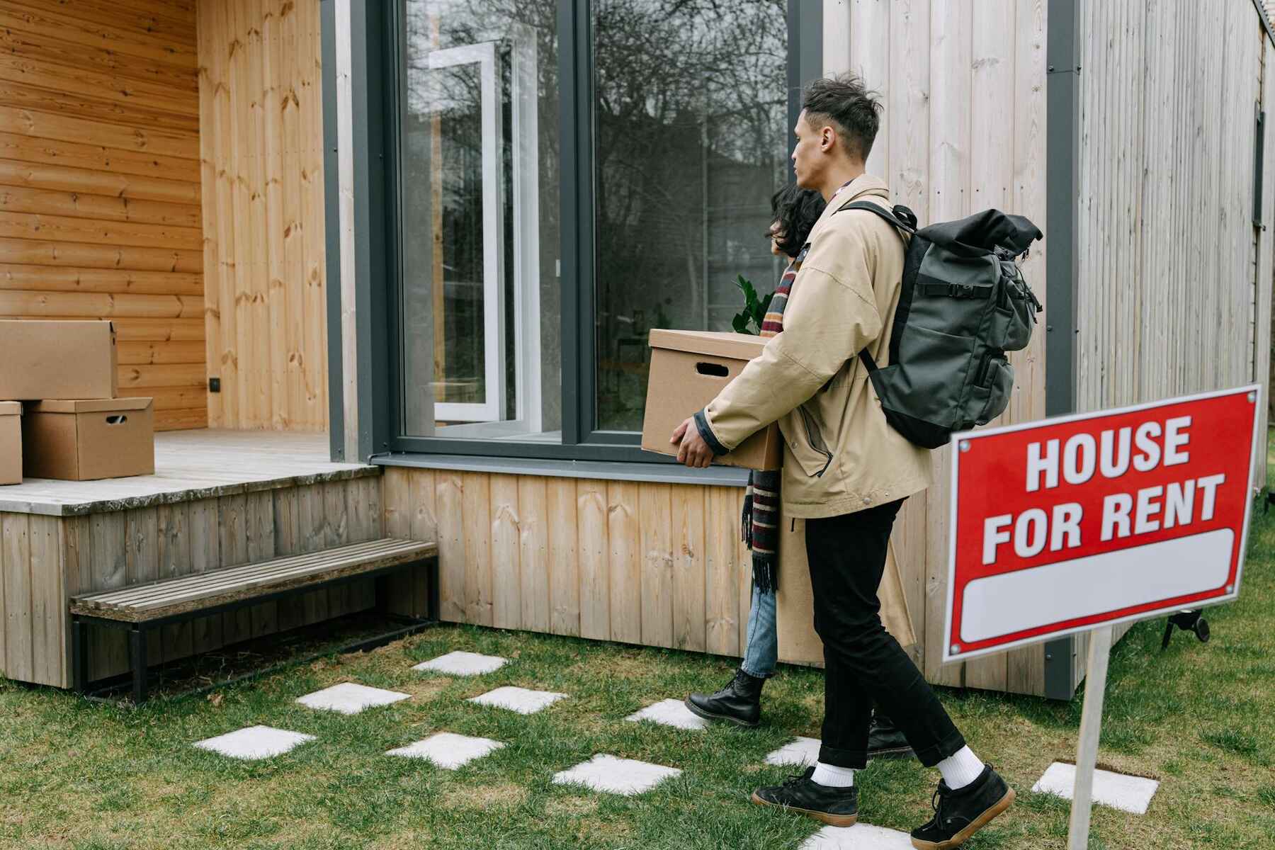 A man and woman hold boxes outside a house with a "For Rent" sign